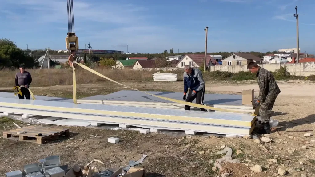 Трое монтажников стропят пакет сэндвич-панелей краном перед подъёмом на каркас