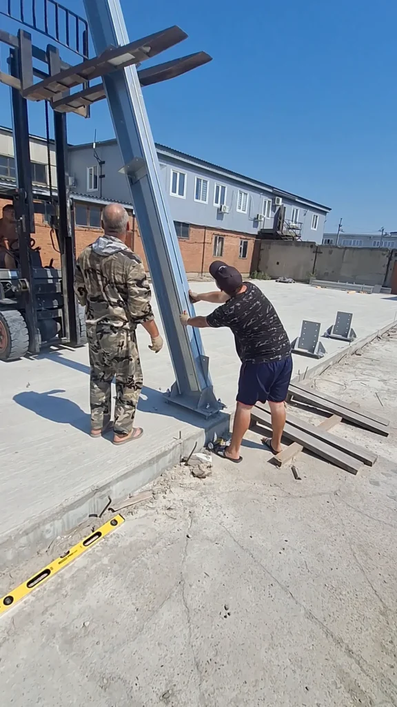 Бригада ставит стальную колонну на анкерную базу, погрузчик страхует стойку