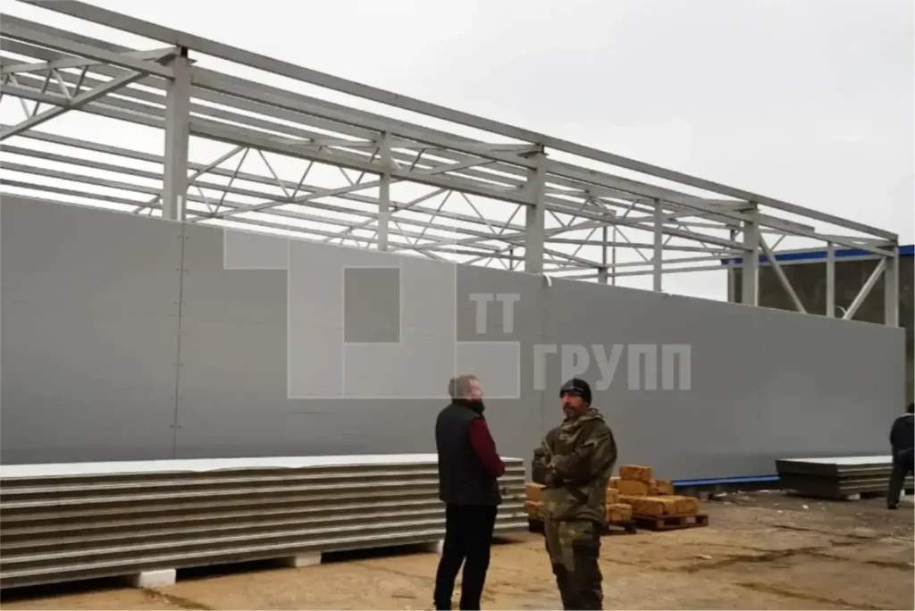 Двое рабочих обсуждают монтаж на фоне зашитой панелями стены, выше открыт стальной каркас с фермами