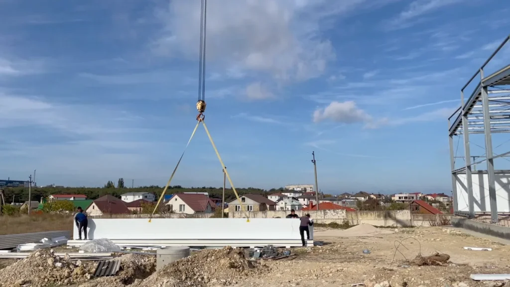Стеновая панель на стропах автокрана у каркаса здания, внизу работают монтажники